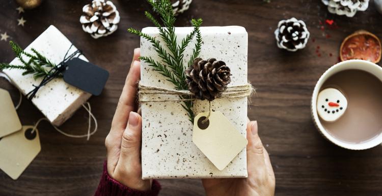 Hands holding wrapped present with pine leaves and a pinecone and hot drink next to it