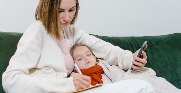 White lady holding on the phone, taking notes, sitting with her sick child.