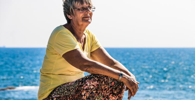 Older white woman sitting on rocks at the beach looking out to sea