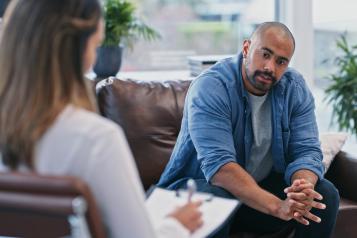 Black man sitting on a sofa talking to a woman who is holding a clipboard