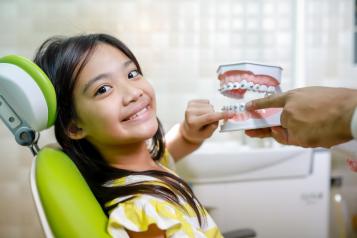 Young Asian girl in a green dentist chair pointing to braces on a model of plastic teeth