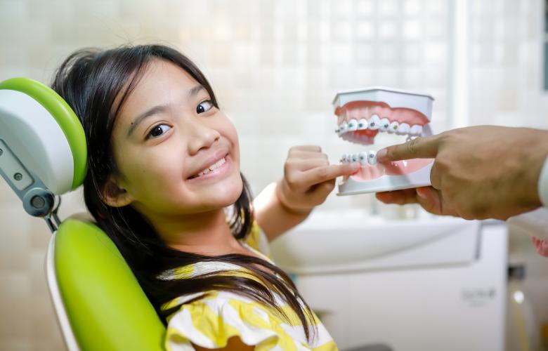 Young Asian girl in a green dentist chair pointing to braces on a model of plastic teeth